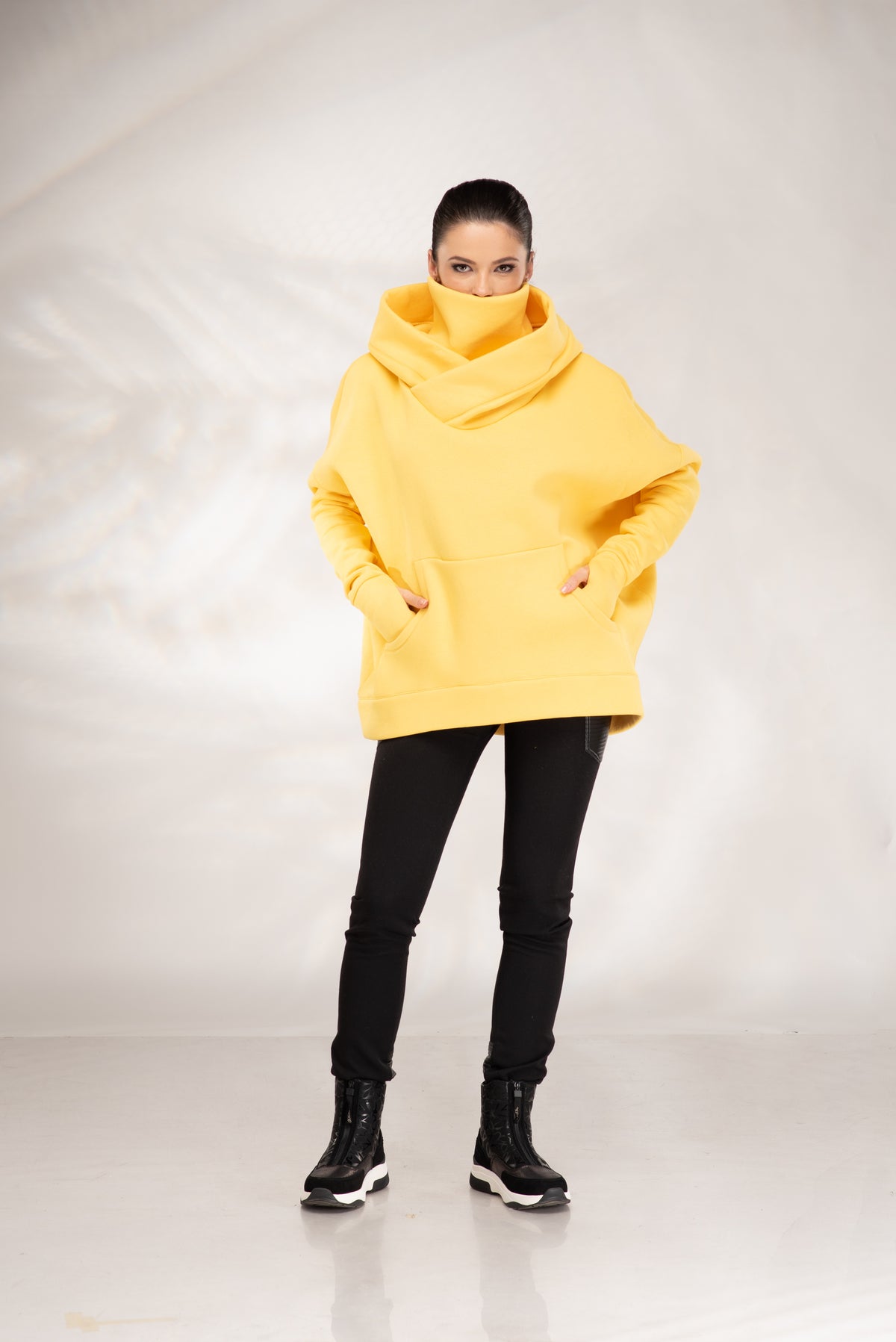 A woman modeling a bright yellow oversized cowl-neck hoodie with front pockets, paired with black pants and black lace-up boots, standing against a light gray studio backdrop.
