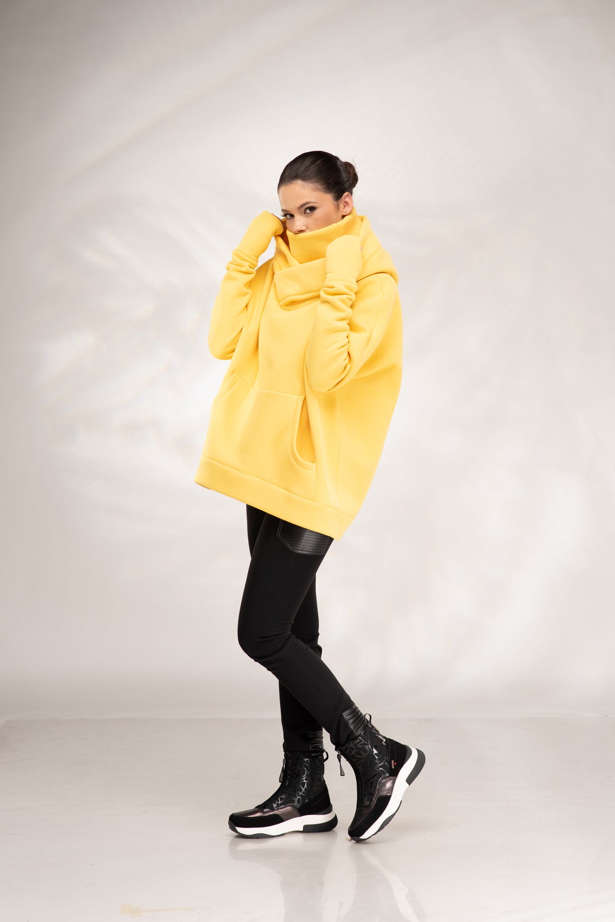 A woman modeling an oversized bright yellow hoodie with a high folded collar, paired with black pants and black sneakers, standing against a light gray studio backdrop.