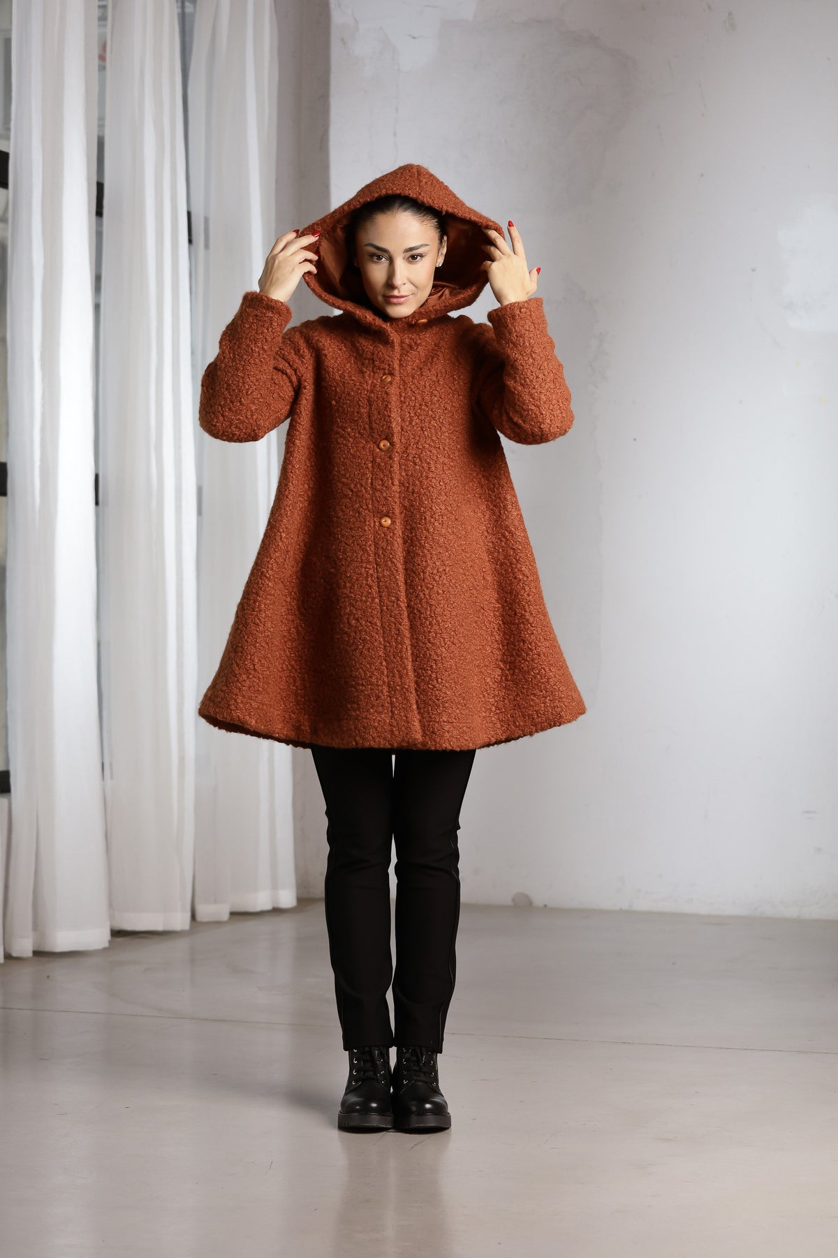 A woman wearing a textured rust-orange hooded coat with buttons, black pants, and black boots, standing in a minimalist studio with white curtains and a light gray wall.