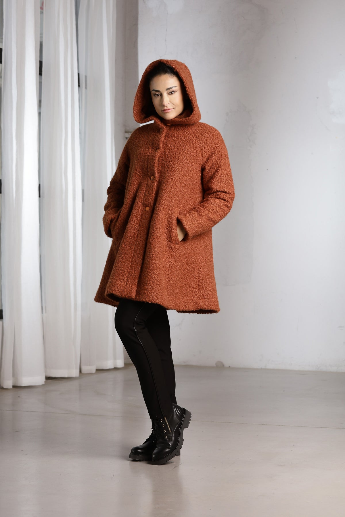 A woman wearing a textured, rust-orange hooded coat with black pants and black boots, standing in a minimalist studio with white curtains and a light gray floor.