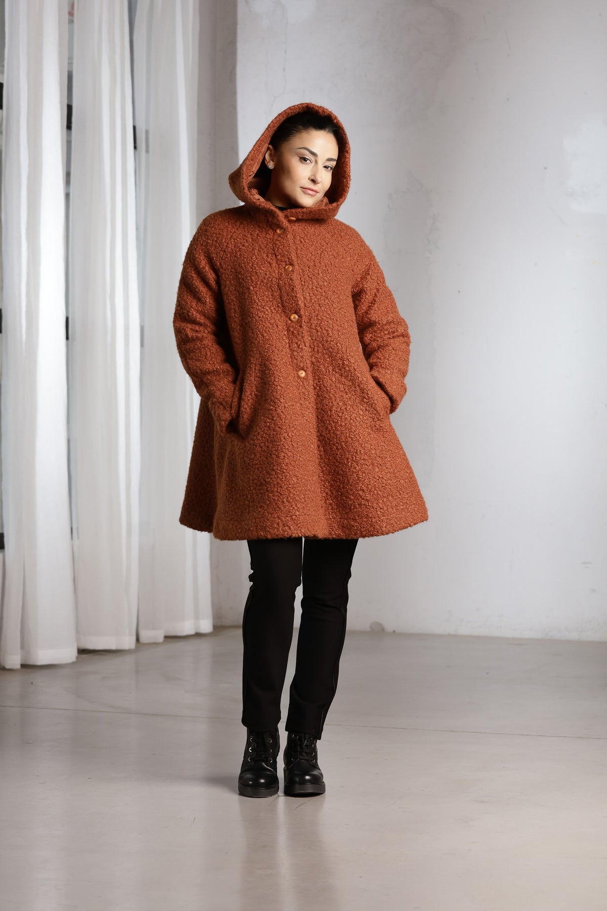 A woman wearing a textured rust-orange hooded coat with buttons, paired with black pants and black boots, standing in a minimalist studio with white curtains and a light gray wall.