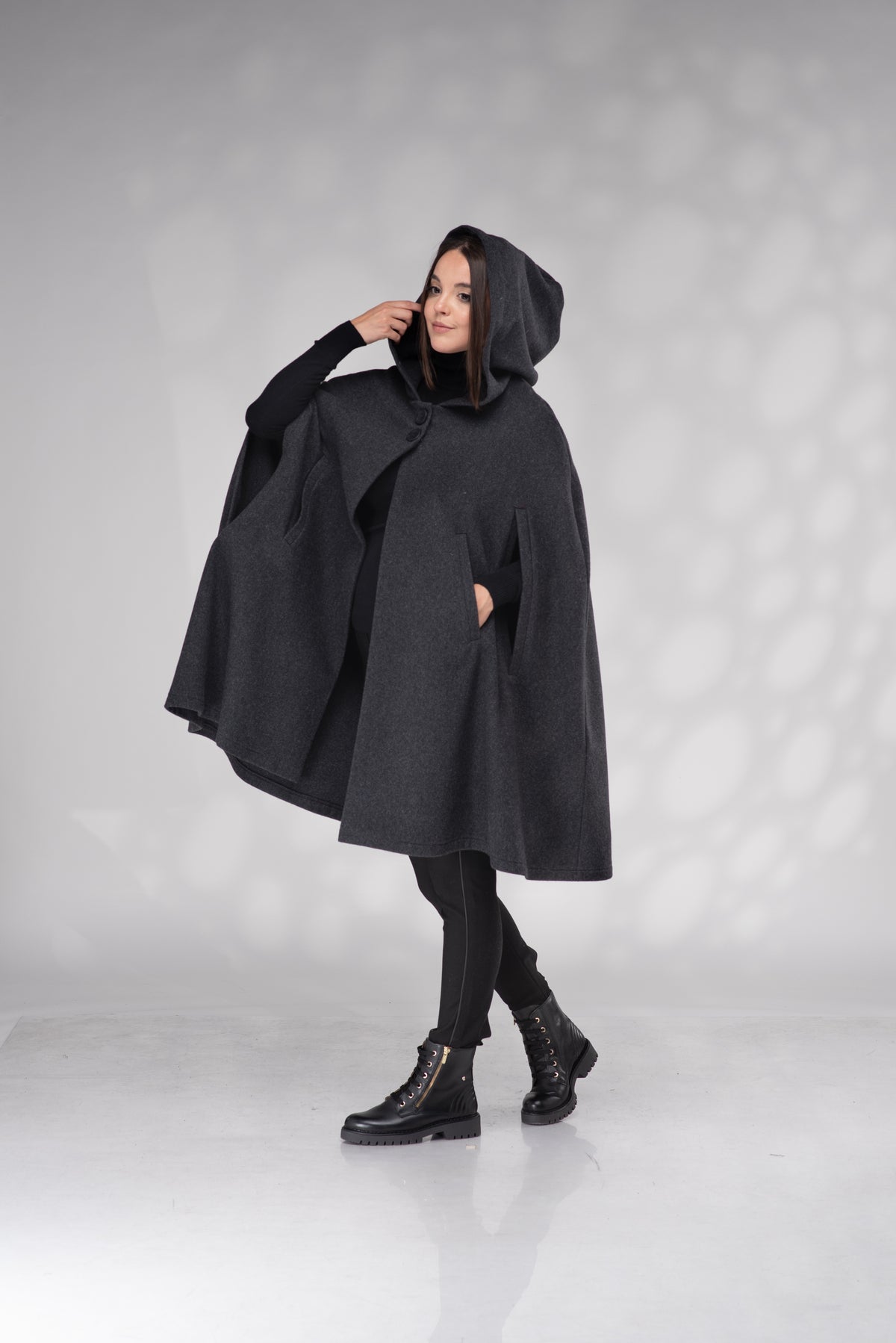 A woman modeling a dark charcoal gray hooded cape with front pockets, styled with black leggings and black lace-up boots against a light gray studio backdrop with soft shadow patterns.